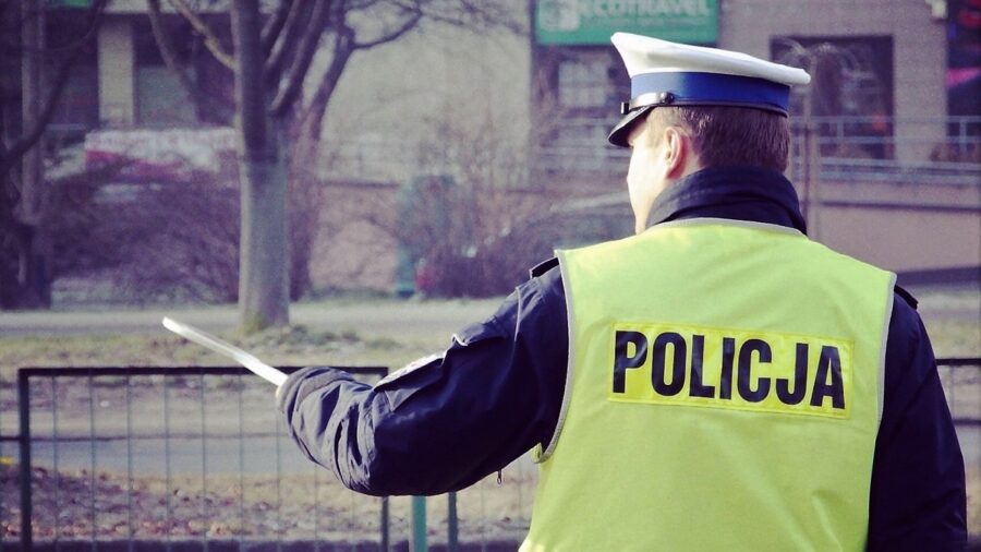 Policjant ruchu drogowego stojący tyłem, ma założona kamizelkę odblaskową z napisem Policja, w lewej ręce trzyma tarcze sygnalizacyjną